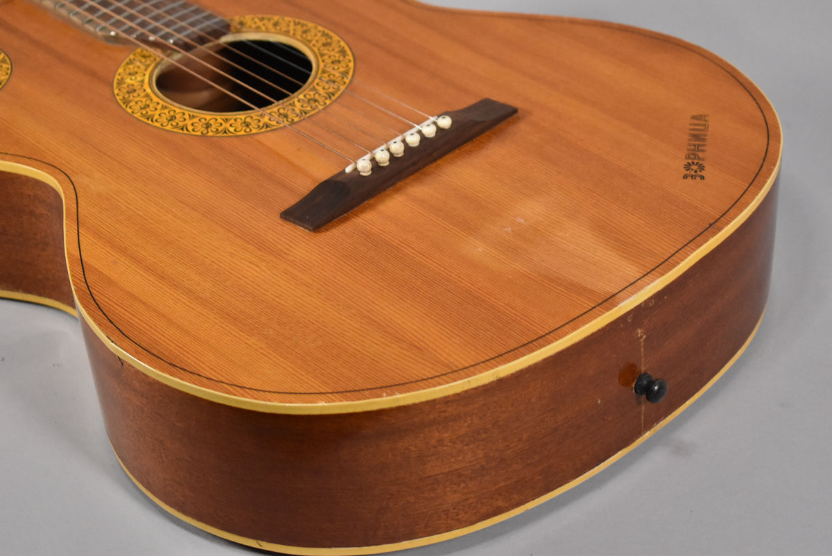The natural finish acoustic guitar's body is angled, showing the ornate soundhole rosette, bridge, and a black strap button.