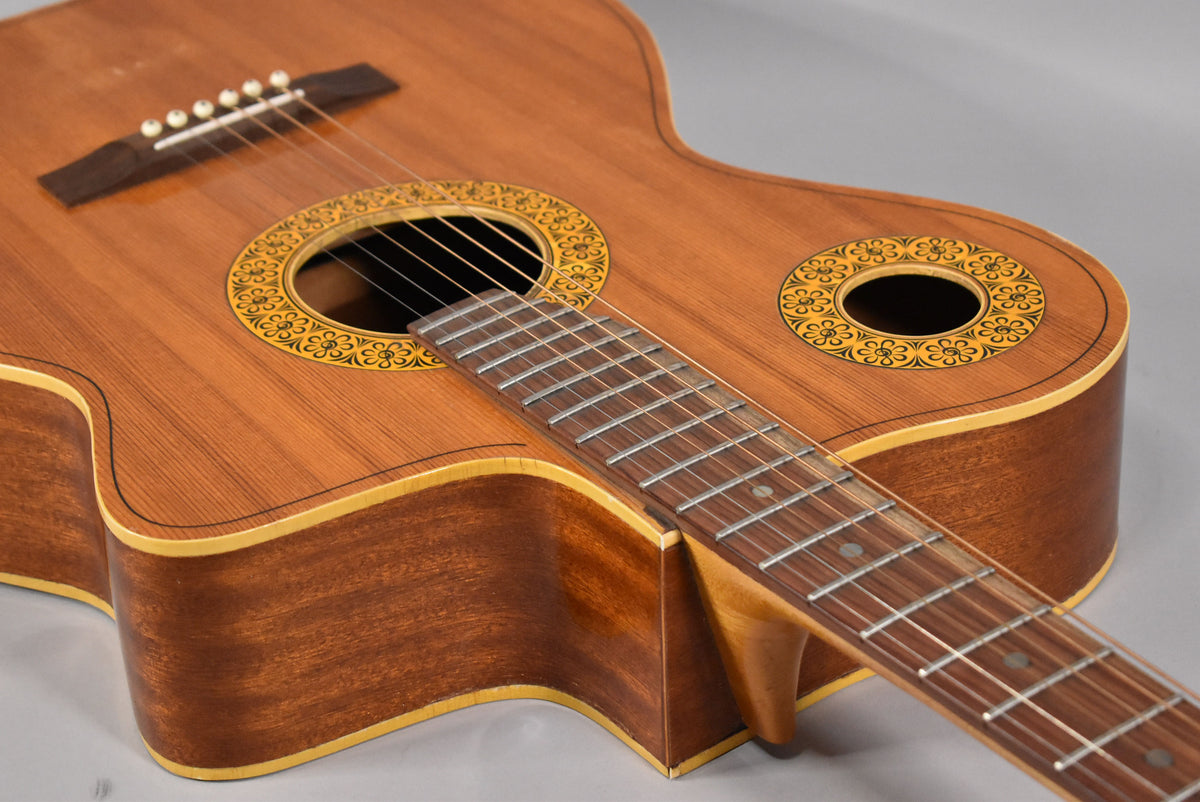 The natural finish acoustic guitar has two soundholes with yellow and black floral rosettes, and a dark fretboard with dot inlays.