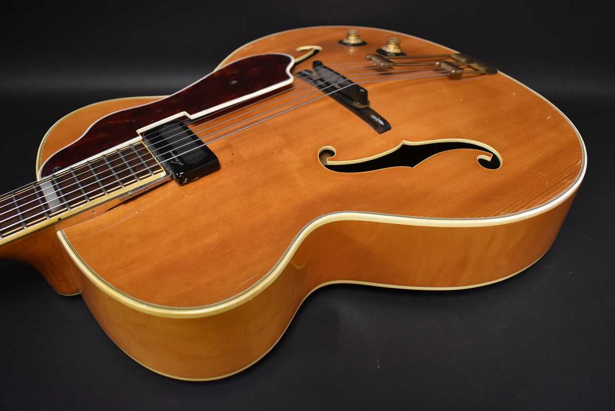 The upper body of a natural finish archtop guitar, showing a dark pickguard, f-holes, and two gold control knobs.