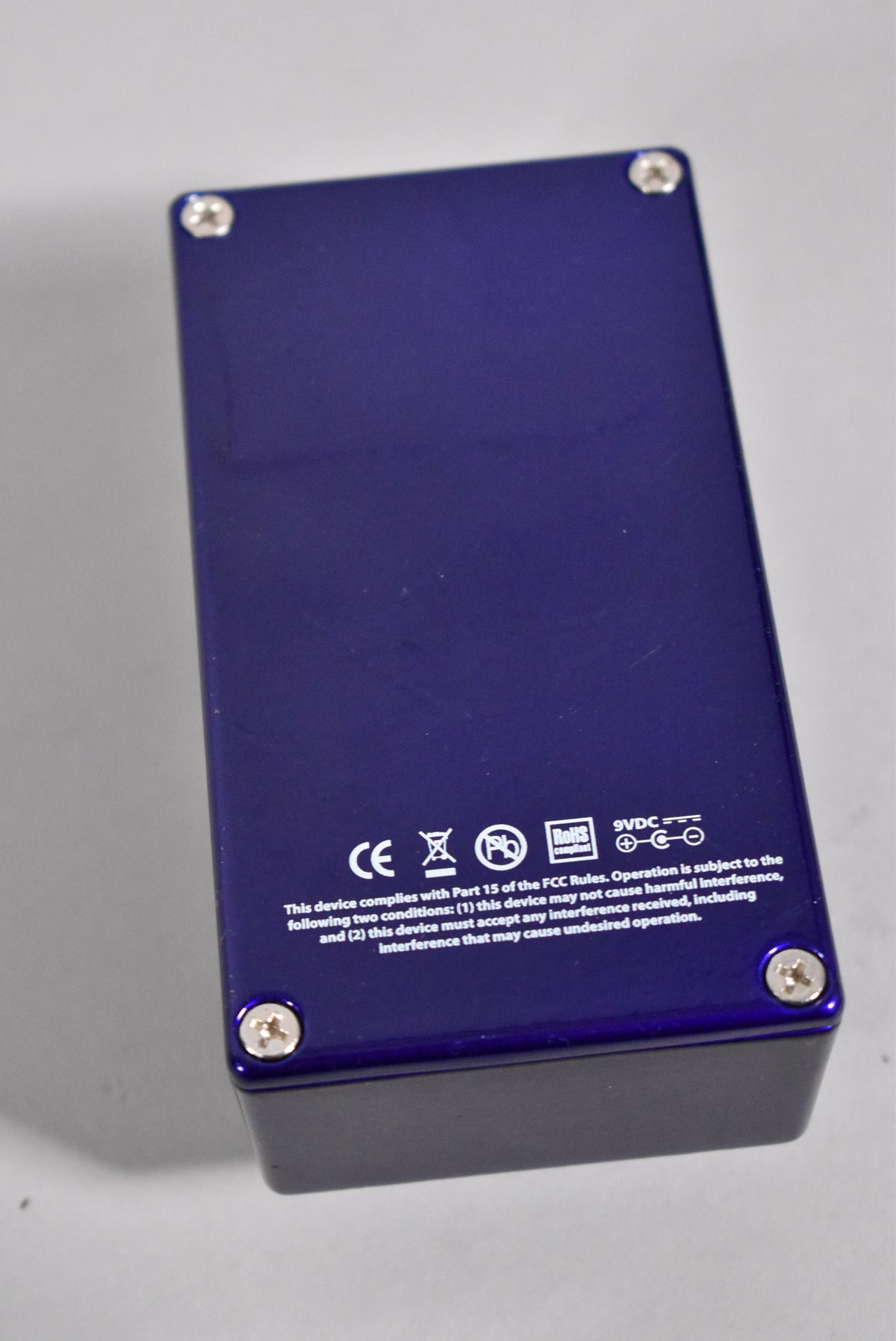 The pedal's dark blue underside is shown at an angle, revealing four silver screws and white regulatory text.