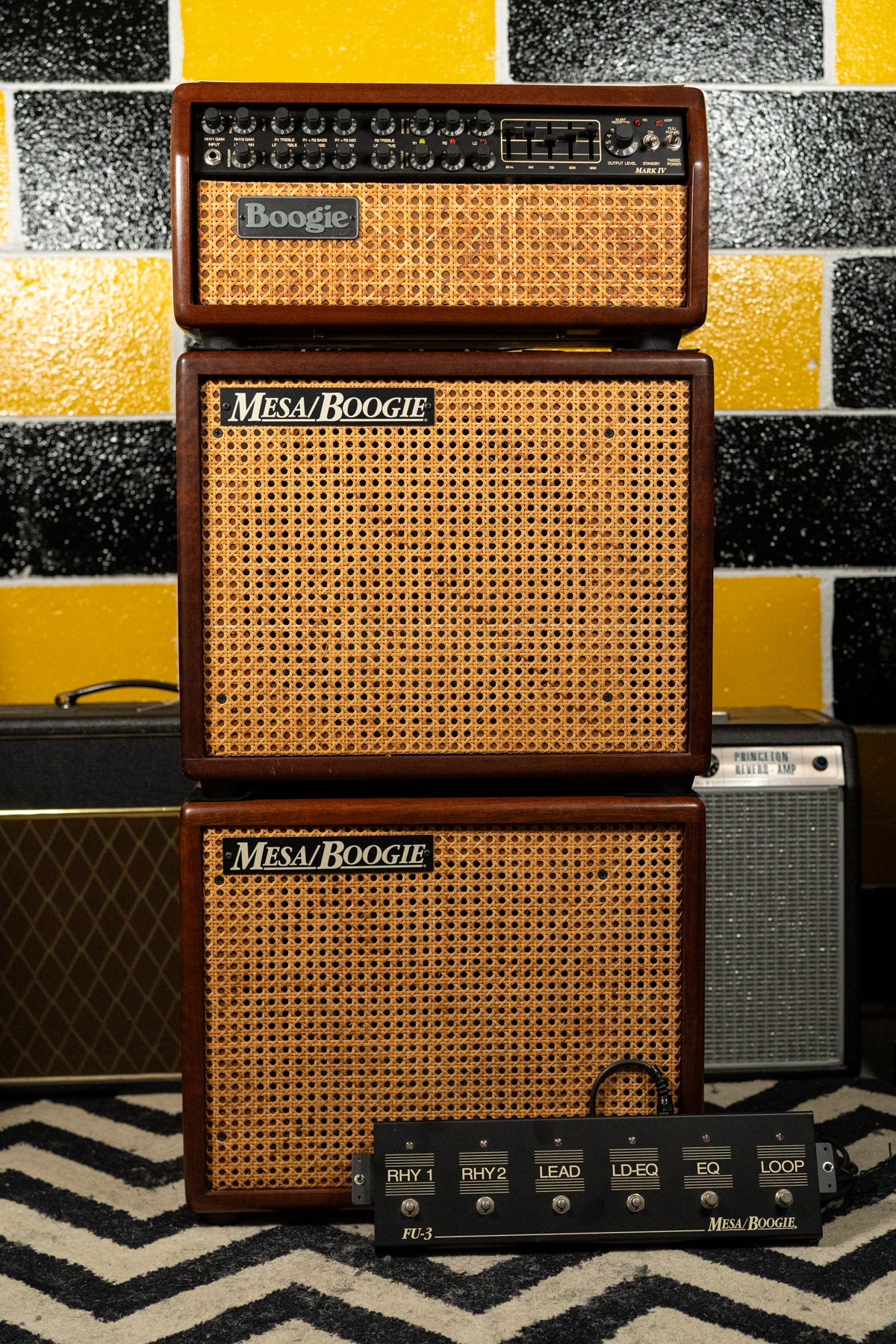 A dark wood amp head with a wicker grille sits atop two matching speaker cabs, with a black footswitch on the rug below.