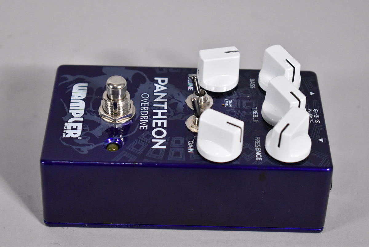 A dark purple pedal with a subtle geometric pattern, five white control knobs, two small toggle switches, and a footswitch.