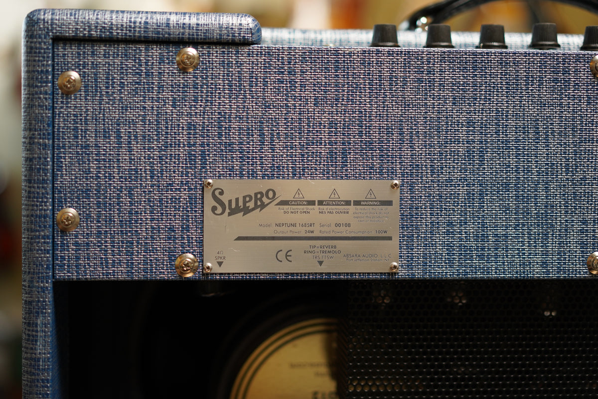 Rear view of the blue tweed amp, showing a silver metal plate with text, four gold screws, and a speaker grille below.