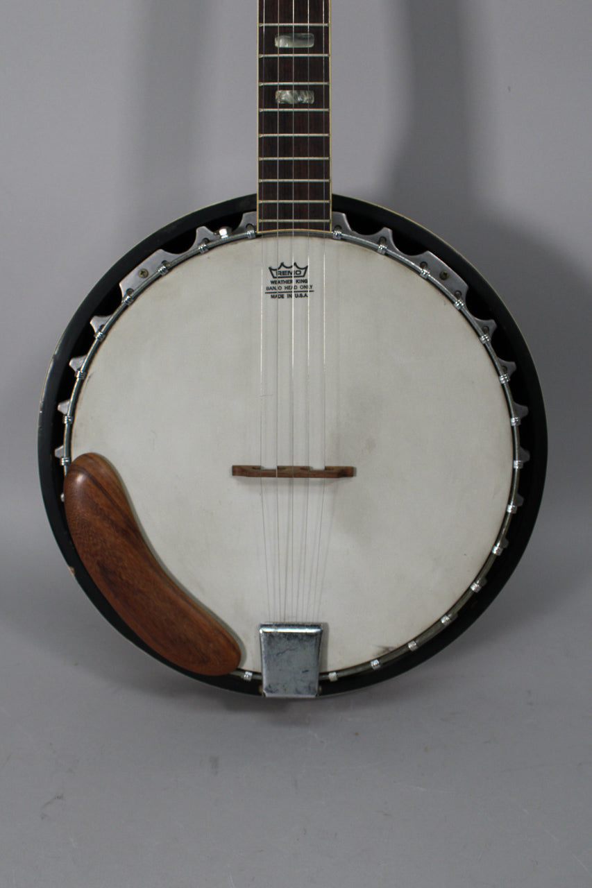 The banjo's white head and dark wood neck with rectangular inlays are framed from the mid-neck to the tailpiece, showing a wooden armrest.