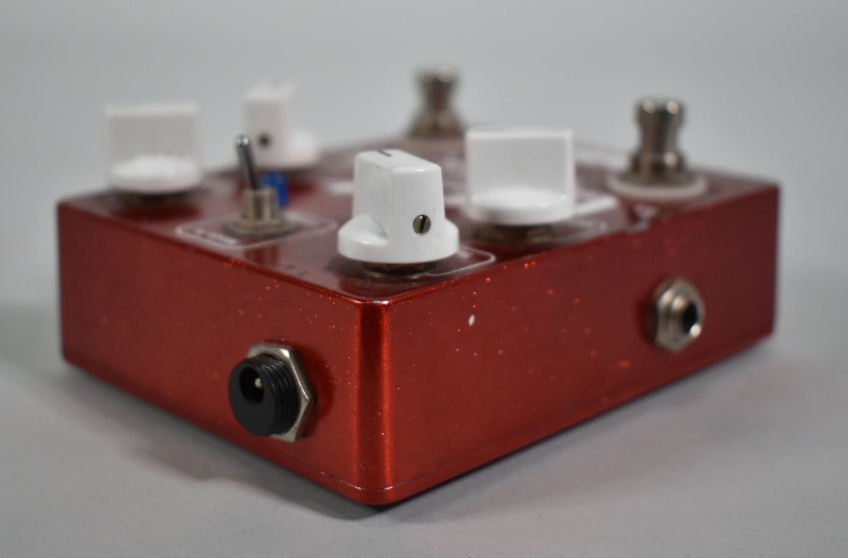 A red glitter-finish pedal, angled to show the left side with a power input and the top with white knobs and a toggle switch.