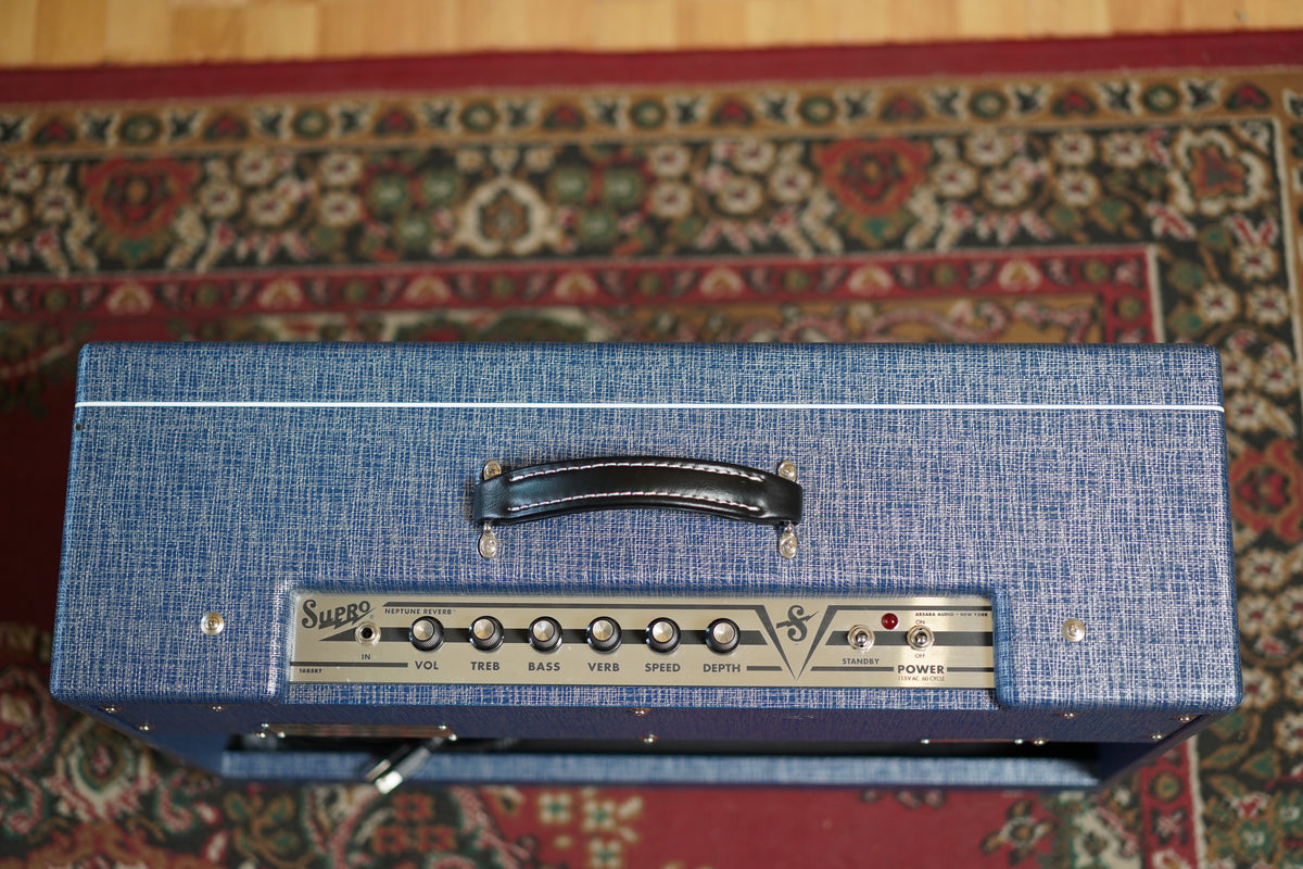 Top-down view of a blue textured amp with a black handle, a gold control panel, and six silver knobs.
