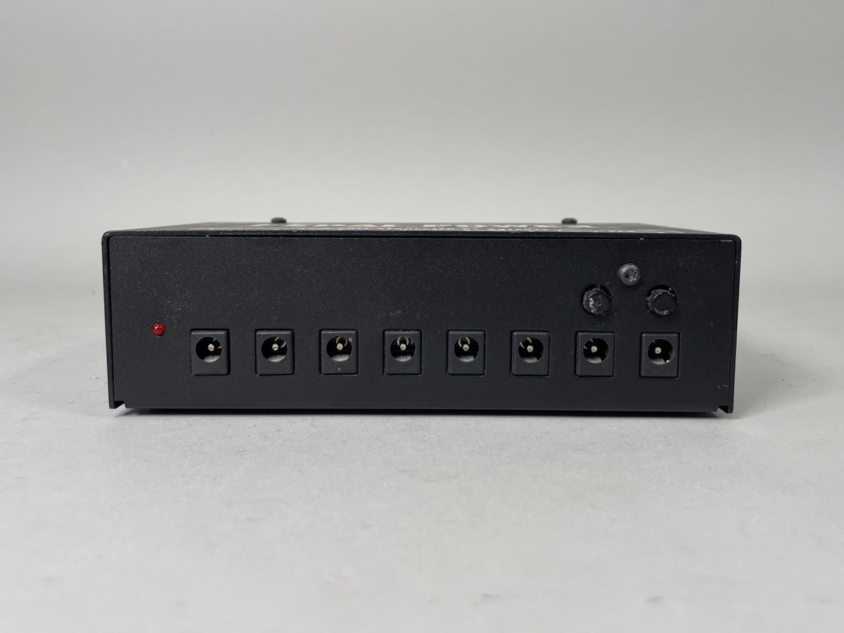 Rear view of the black pedal power supply, showing eight DC outputs, a red indicator light, and two worn black control knobs.