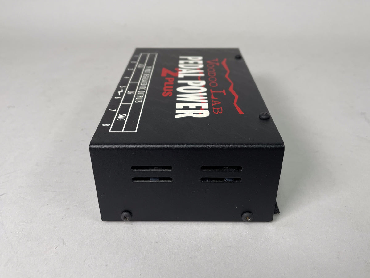 A black rectangular pedal power supply is angled right, showing its top with white and red lettering, and its vented right side.