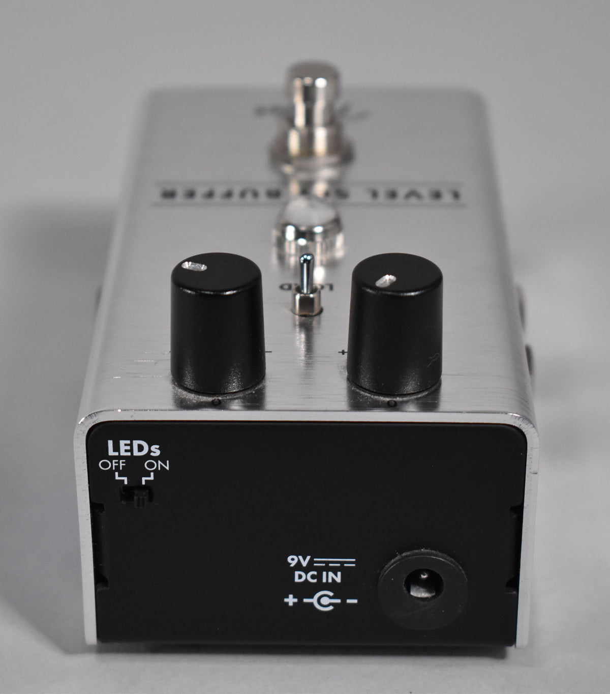 A brushed silver pedal with black front panel, showing two black knobs, a toggle switch, and a 9V DC input.