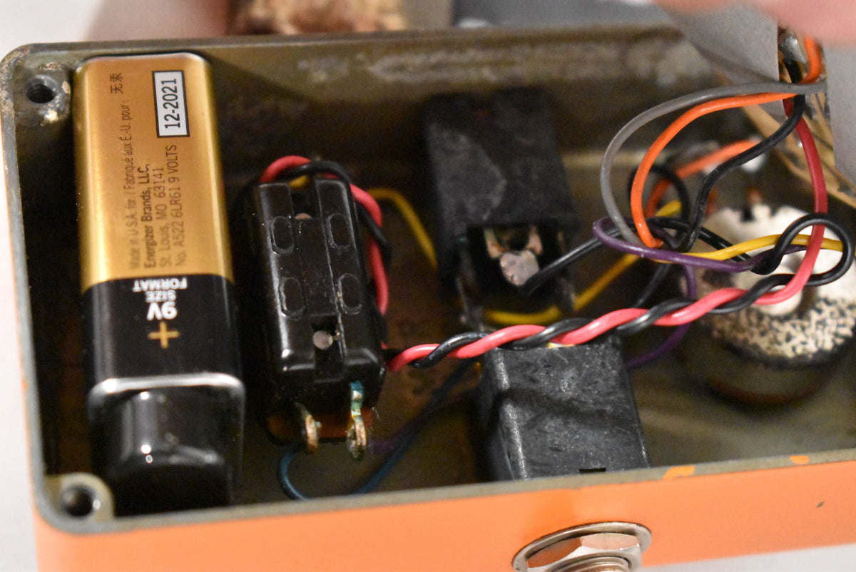 The pedal's orange casing is open, revealing a gold 9V battery, black battery clip, and colorful wiring inside.