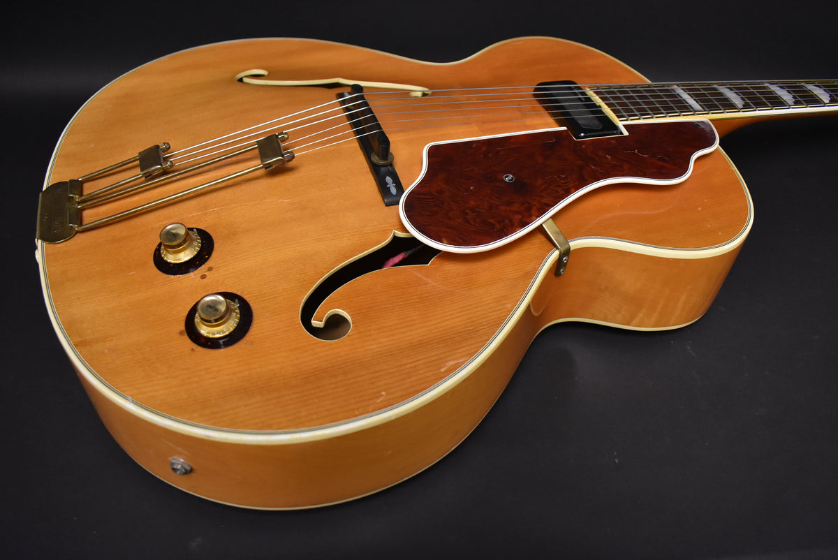 Upper body of a natural finish archtop guitar, showing two gold knobs, a dark tortoise pickguard, and f-holes.