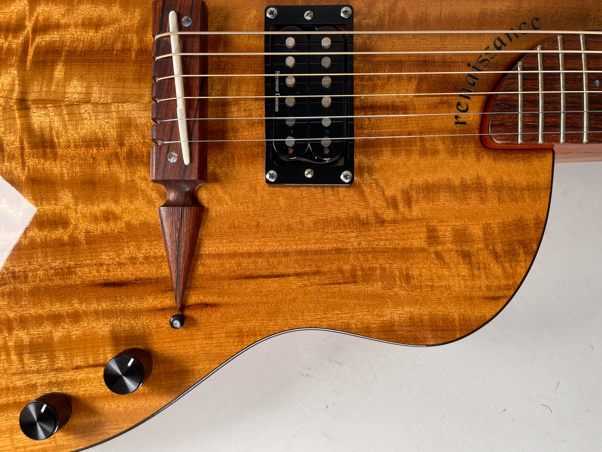 Close-up of the koa guitar body, showing the dark wood bridge, black humbucker pickup, and two black control knobs.