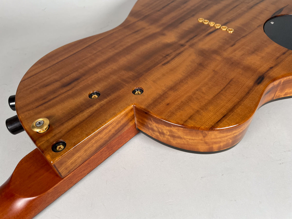 The guitar's glossy, figured koa back is angled, showing the neck joint, gold string ferrules, and a black control plate.