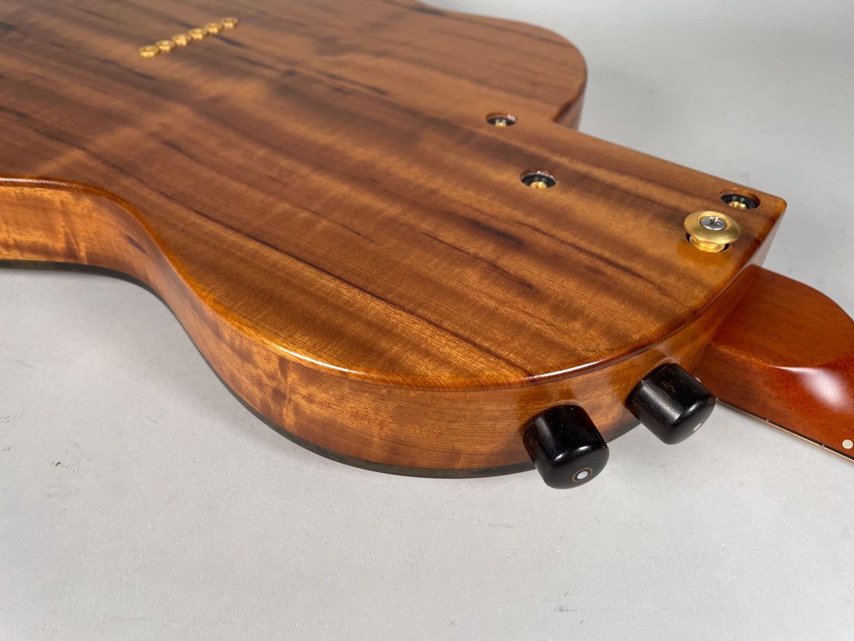 The guitar's glossy, figured koa back is angled, showing two black control knobs and a gold output jack on the lower bout.