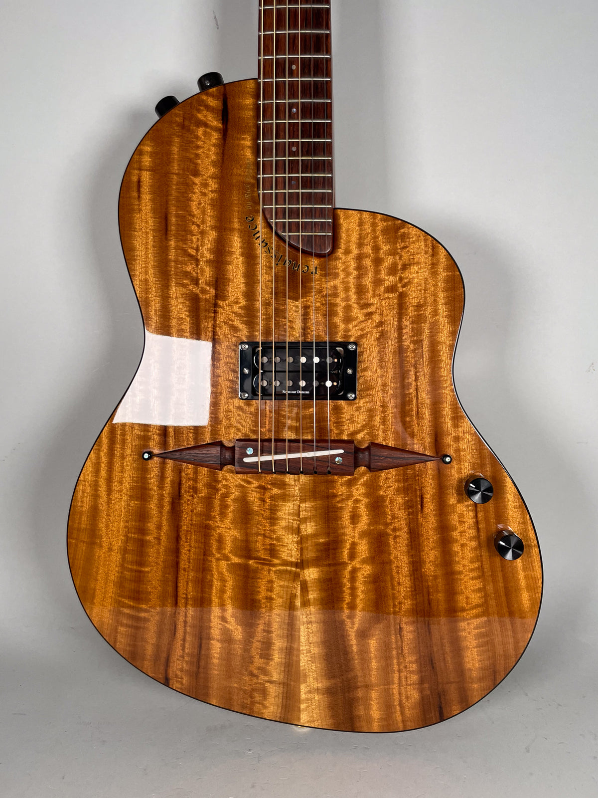 The guitar's glossy, figured koa body is framed from the neck joint to the lower bout, showing a black humbucker and two control knobs.