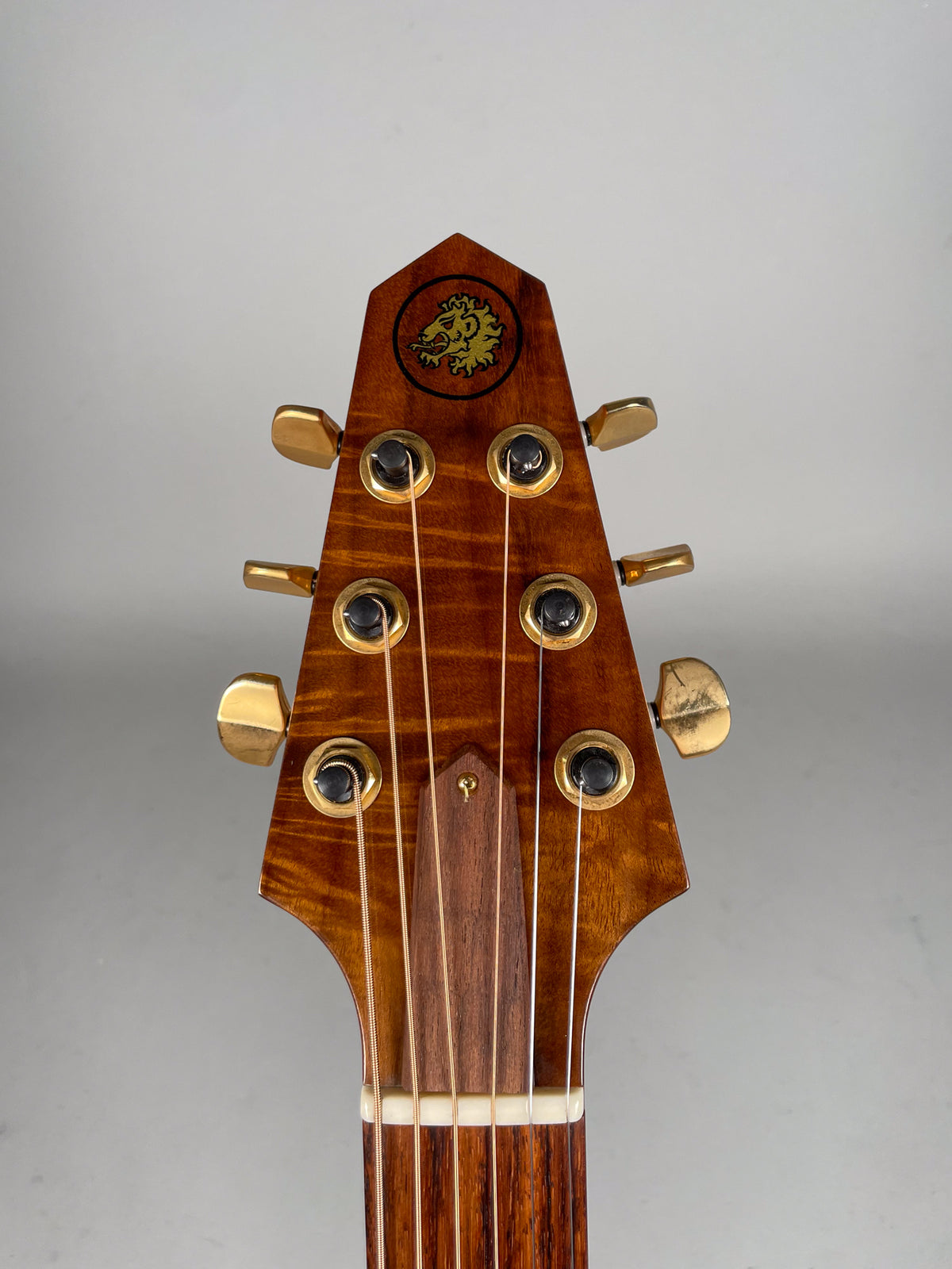 Close-up of the guitar's headstock, showing the figured koa wood, gold tuners, and a circular lion logo.