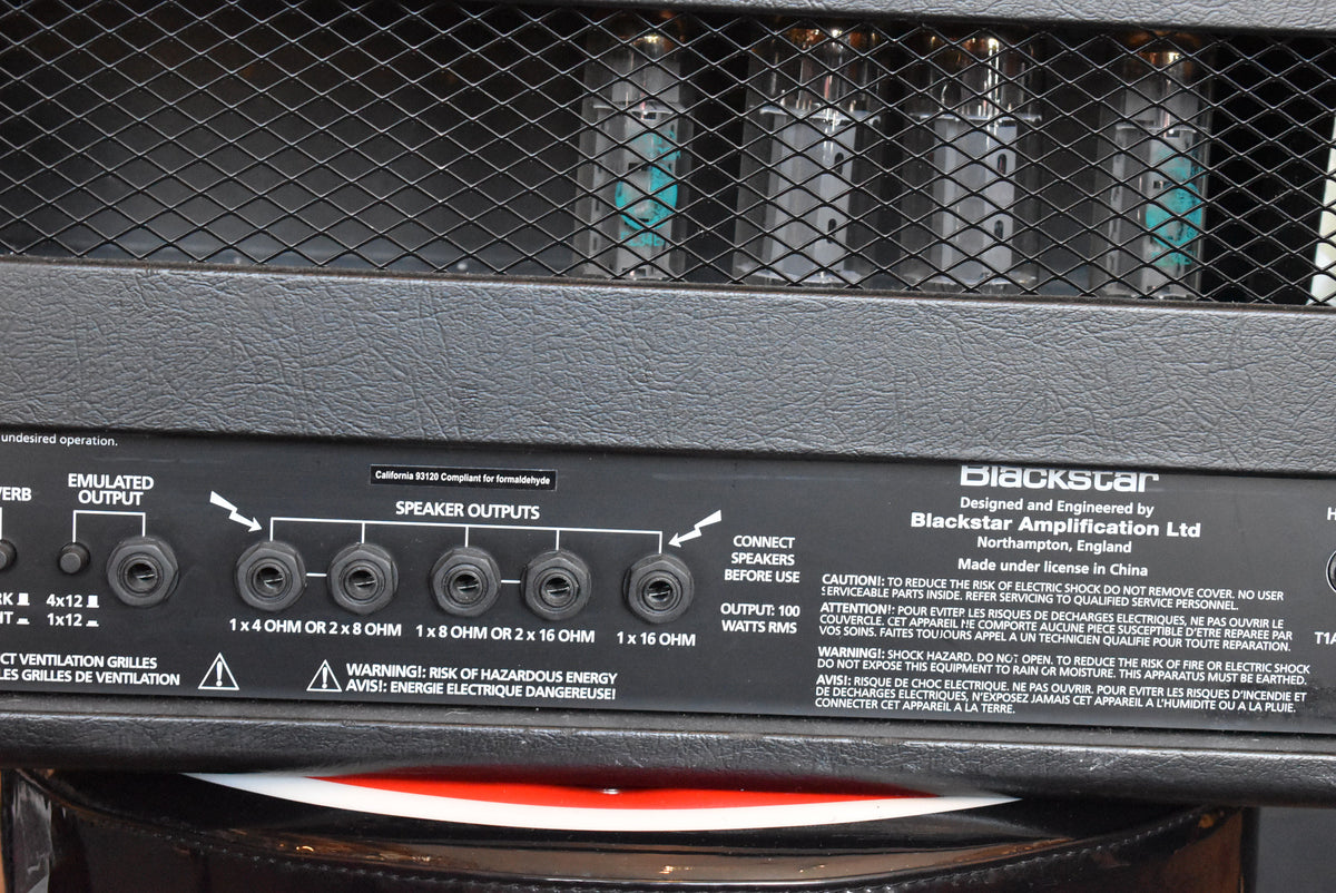 Close-up of the amp's textured black rear panel, showing the speaker outputs and a metal mesh revealing four glowing blue vacuum tubes.