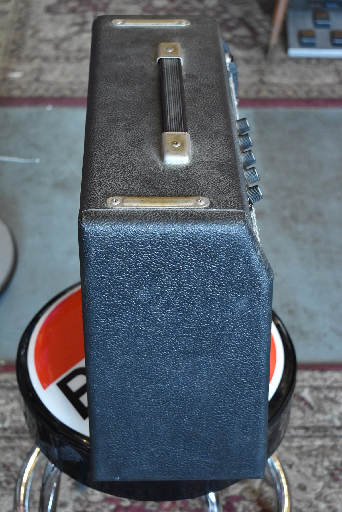 Top-down view of a dark gray textured amp with a black handle, four silver knobs on the right, and some visible wear on the edges.