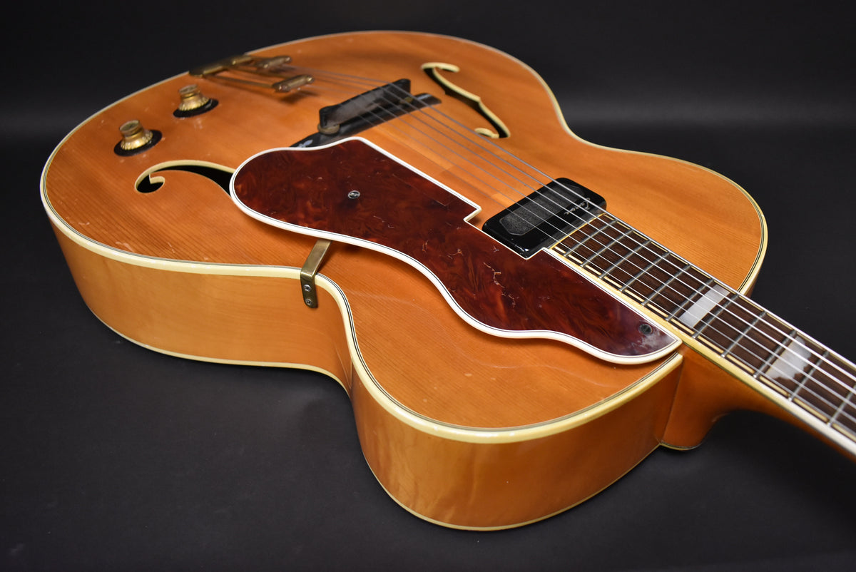 Upper body of a natural finish archtop guitar, showing two gold knobs, f-holes, a dark tortoise pickguard, and neck pickup.