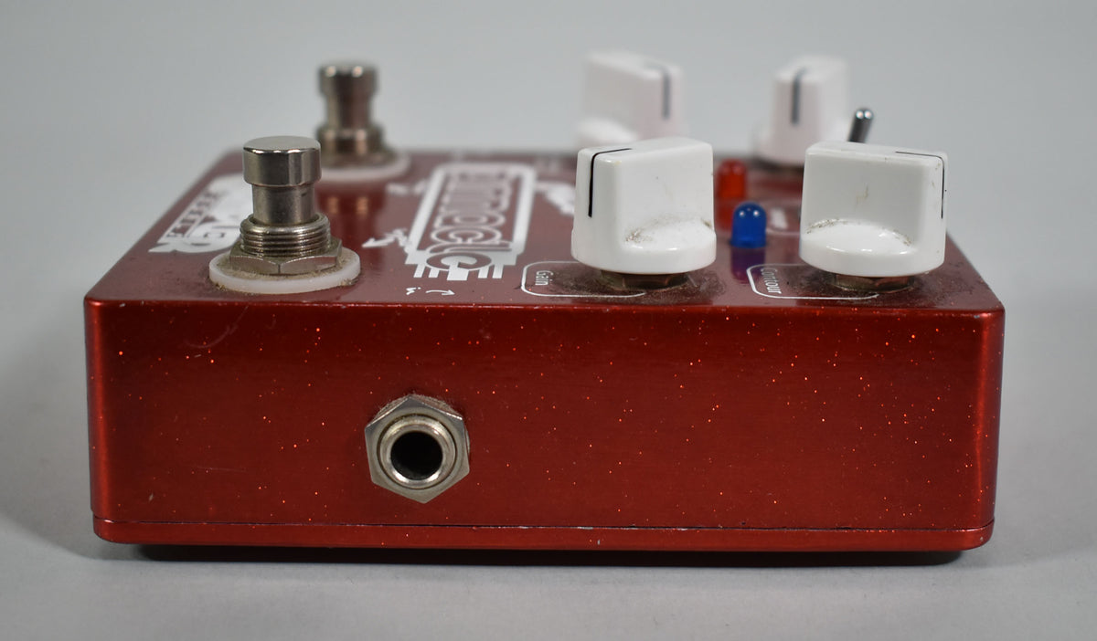 The red glitter-finish pedal is viewed from the side, showing a silver input jack, two footswitches, and three white knobs.