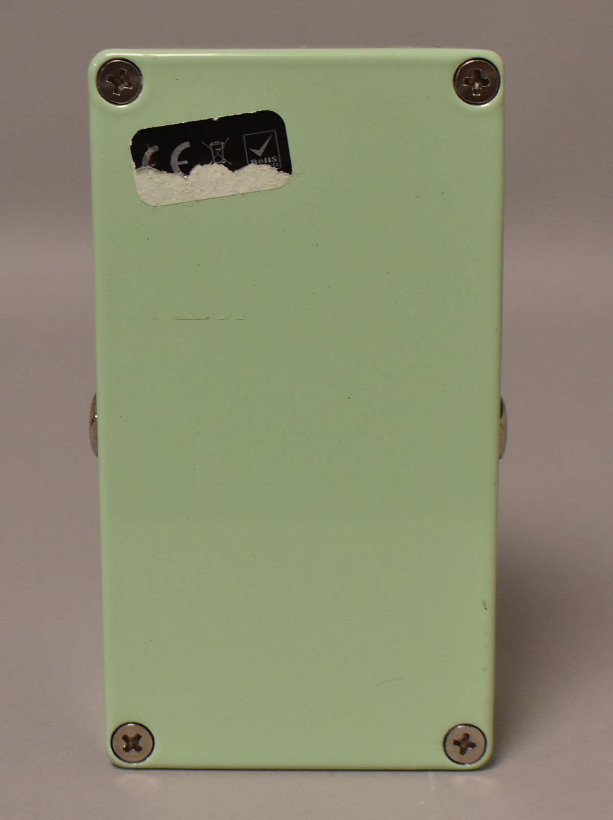 The bottom of the green effect pedal is shown, secured by four silver Phillips head screws, with a partially torn white sticker in the top left.