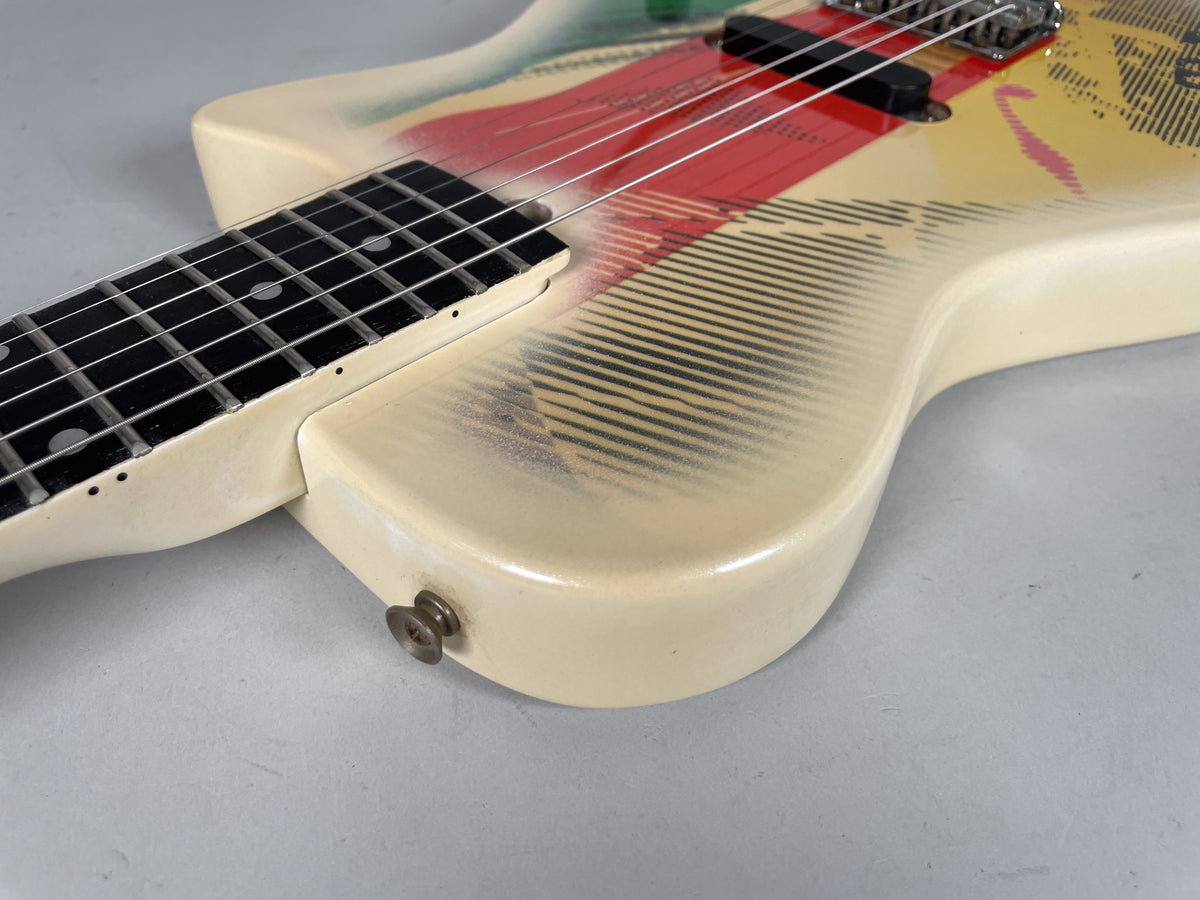 Close-up of the guitar body's lower bout, showing the cream finish with red, yellow, and black graphics, and a strap button.