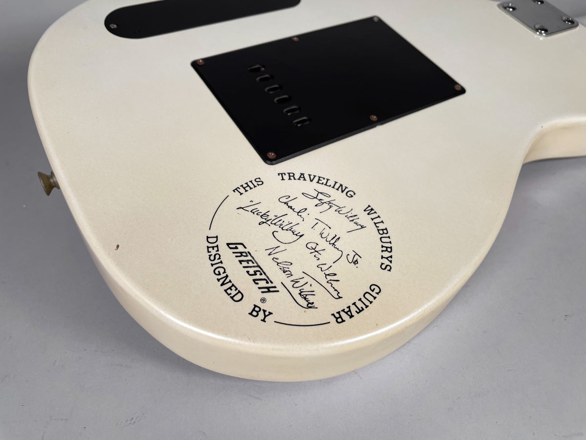 The back of a cream-colored electric guitar, showing a black tremolo cover and a circular graphic with signatures.