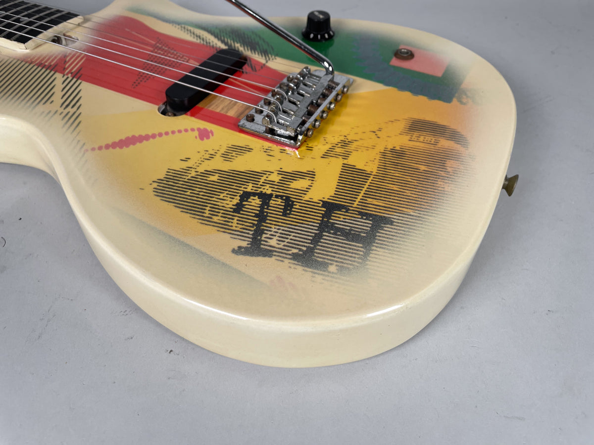 Close-up of the cream-bodied guitar, showing the vibrato bridge, single black pickup, and colorful graphic with black "TH" lettering.