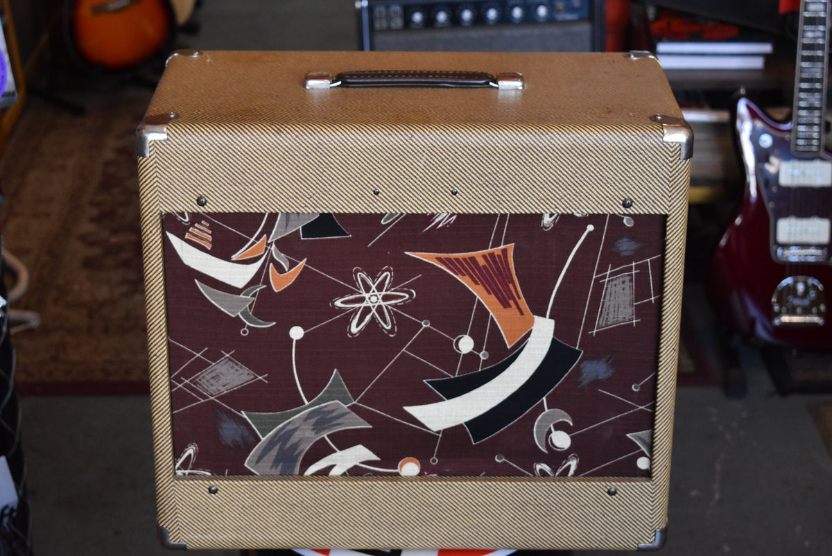Front view of a tweed-covered cabinet with chrome corner protectors, a black leather handle, and a brown grille cloth with an atomic-style pattern.
