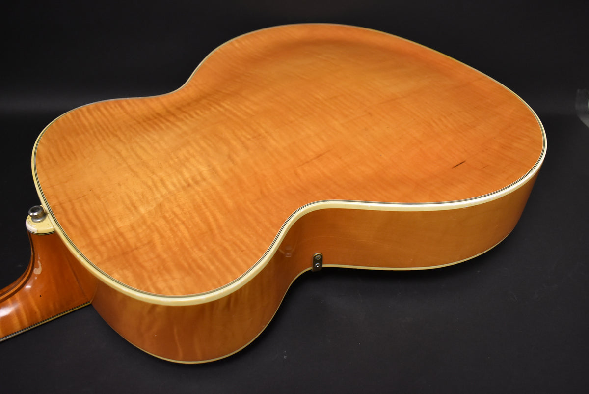The archtop guitar's figured maple back and sides are visible from the lower bout, with a strap button near the cutaway.