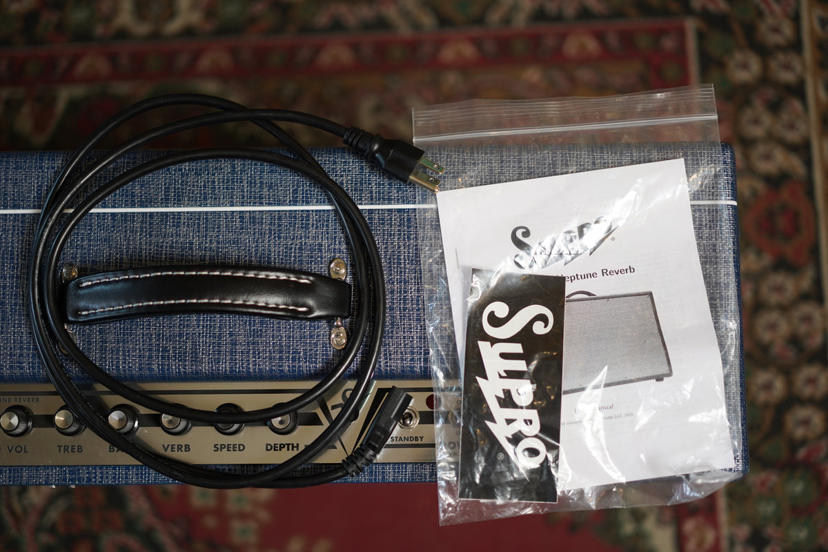 Top-down view of the blue tweed amp, showing the black leather handle, coiled power cable, and sealed bag of paperwork and stickers.