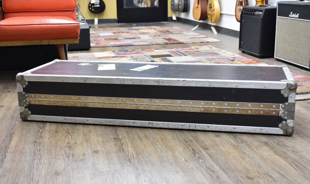 A black flight case with metal trim and corner protectors sits on a wood floor in a guitar shop.
