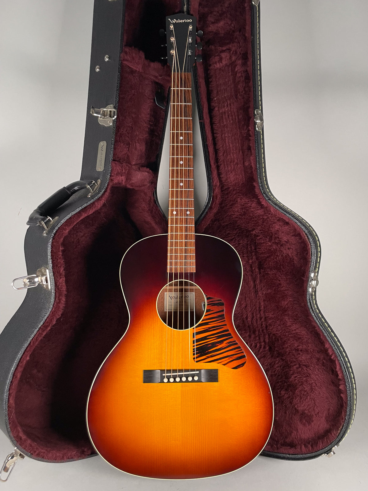 Full-front view of a sunburst acoustic guitar with a tiger-stripe pickguard, standing in its open, dark red-lined hard case.