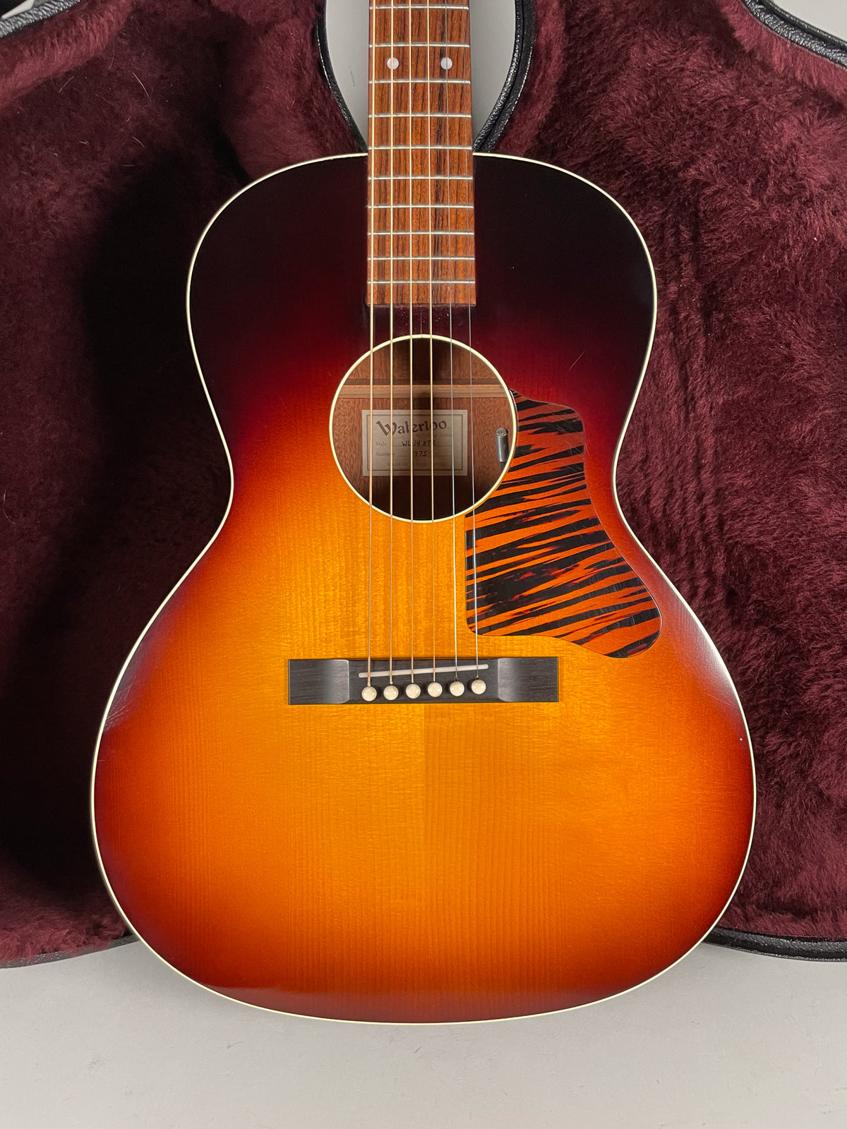 Full-frame view of a sunburst acoustic guitar with a tiger-stripe pickguard, dark bridge, and light fretboard against a burgundy case.
