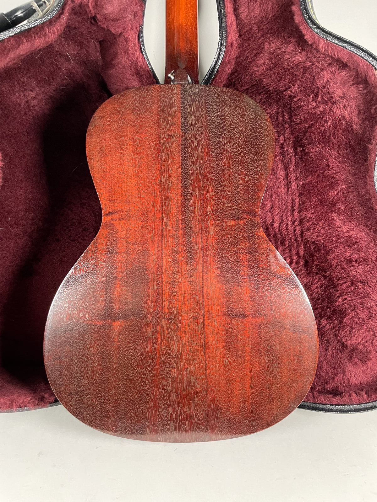 The guitar's dark red-brown back and neck are visible, with a subtle wood grain and a few light scuffs, resting in a red plush case.