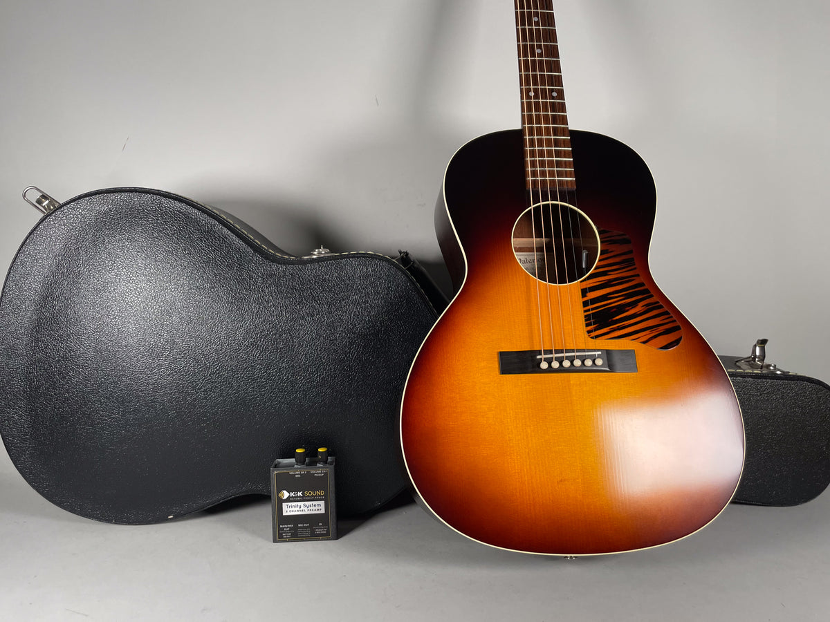 The sunburst acoustic guitar stands upright, angled left, with a black hard case and K&K preamp pedal on the floor.
