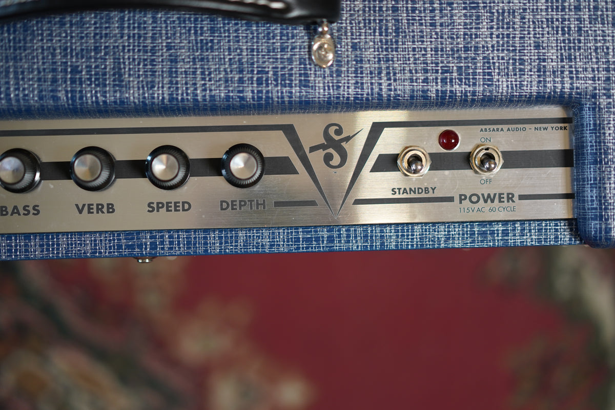 Close-up of the blue tweed amp's silver control panel, showing four knobs labeled Bass, Verb, Speed, Depth, and two toggle switches.