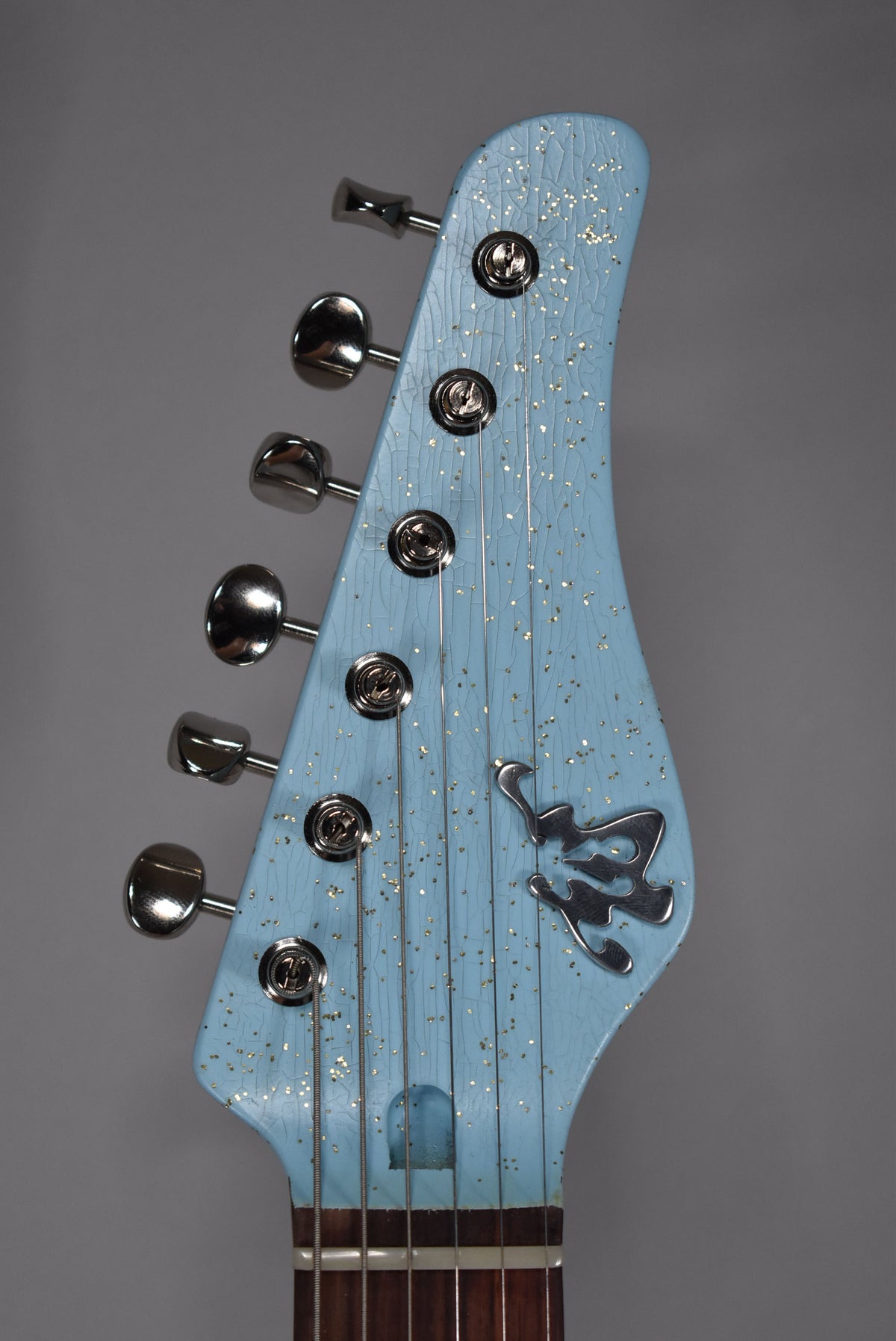 Close-up of the guitar's headstock, showing a sonic blue finish with gold sparkle and relic'd crackle, plus six chrome tuners.
