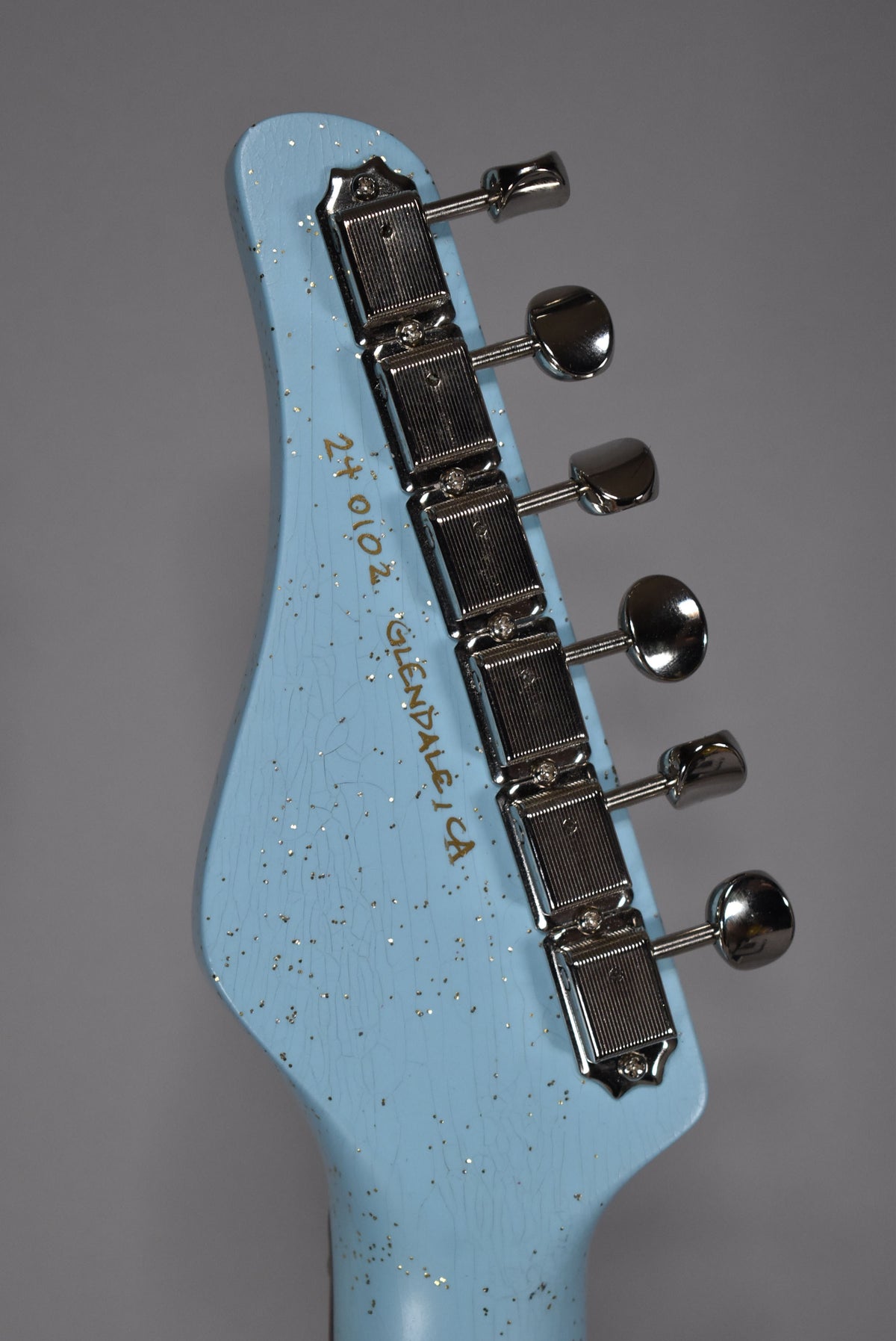 The light blue, glitter-flecked headstock, angled from the left, shows six chrome tuners and fine finish checking.