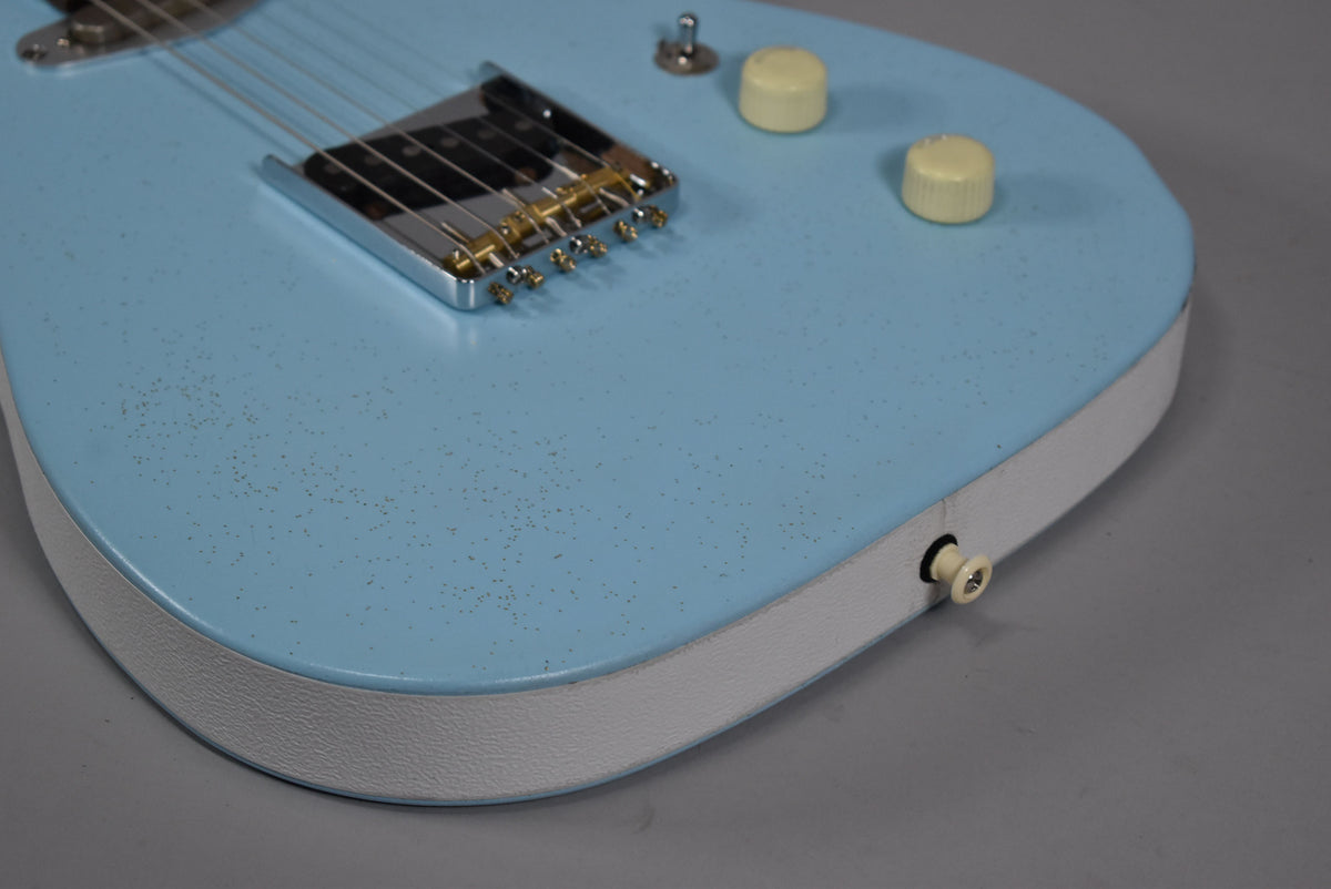 Close-up of the light blue guitar body with a white textured side, showing the chrome bridge, two cream control knobs, and output jack.