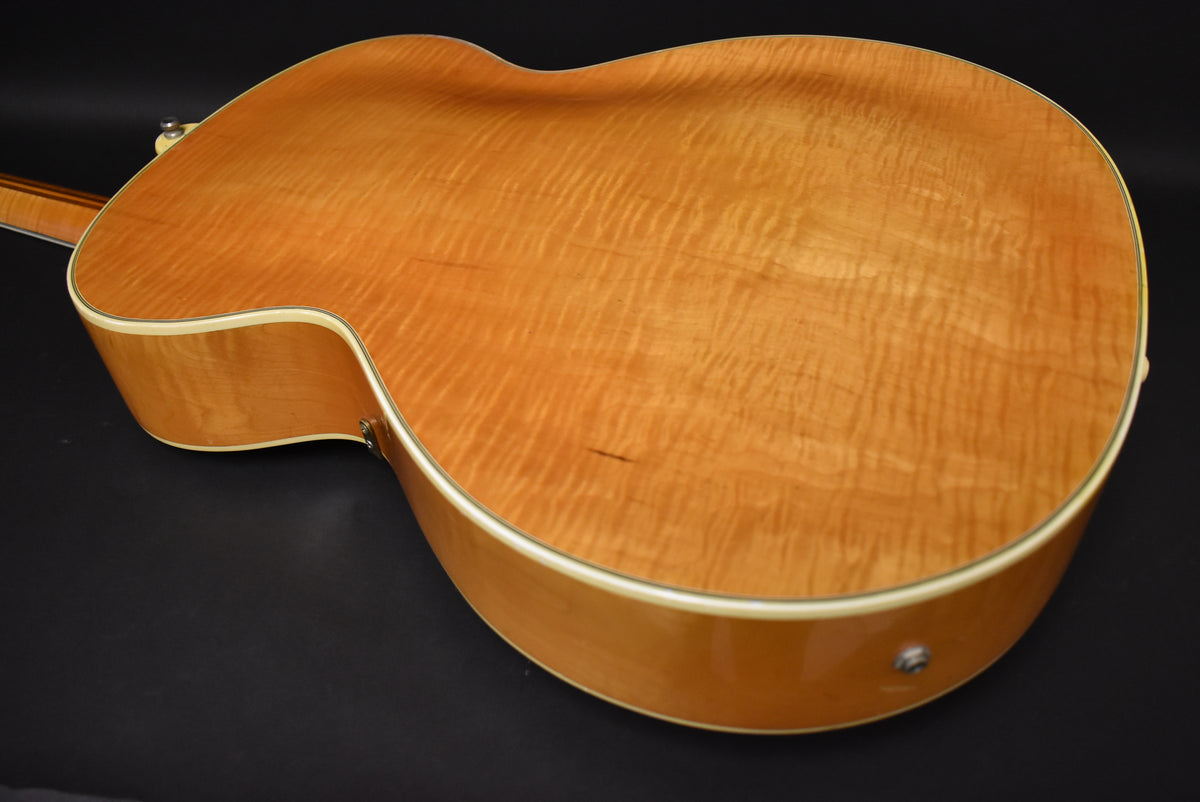 The guitar's back and side are visible, showing a natural finish with prominent flame maple grain and a small dark scratch.