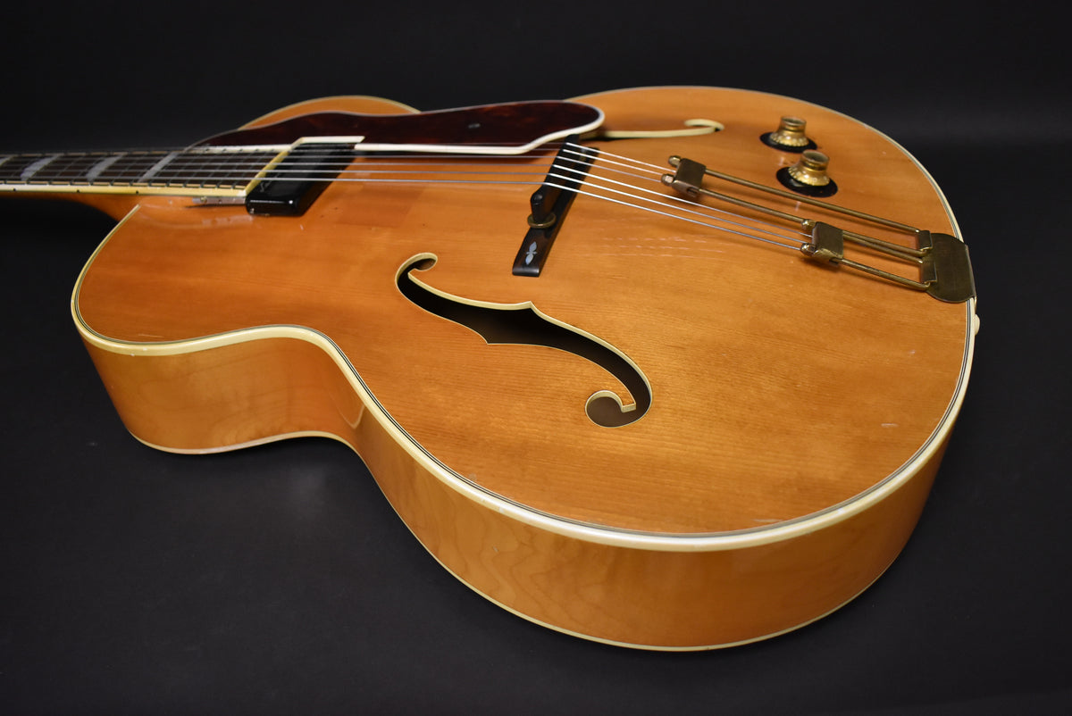 The natural finish archtop guitar is angled from the upper left, showing its light wood body, f-holes, and gold hardware with some wear.