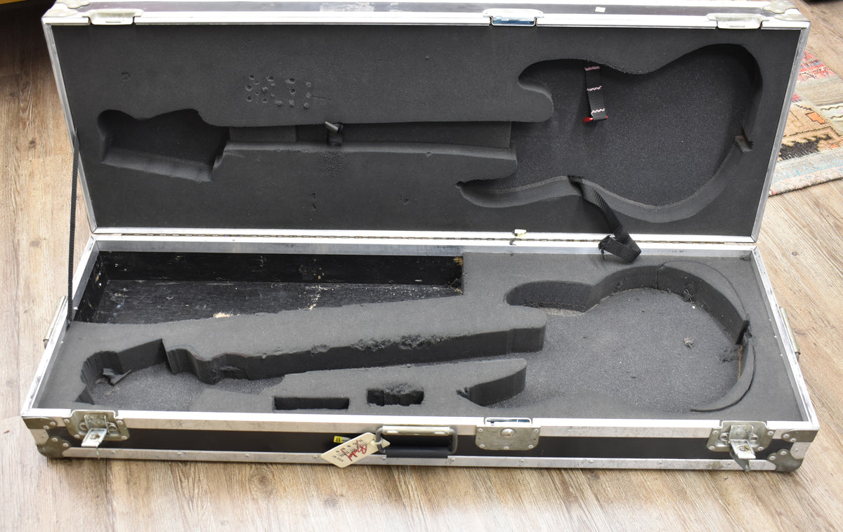 Open black flight case with silver trim, showing custom-cut foam interior for a bass guitar, with visible wear and dust.