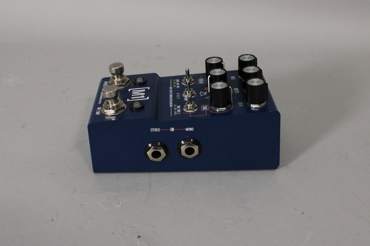 A dark blue guitar pedal, angled from the left, shows two footswitches, six black knobs, three toggle switches, and two input jacks.