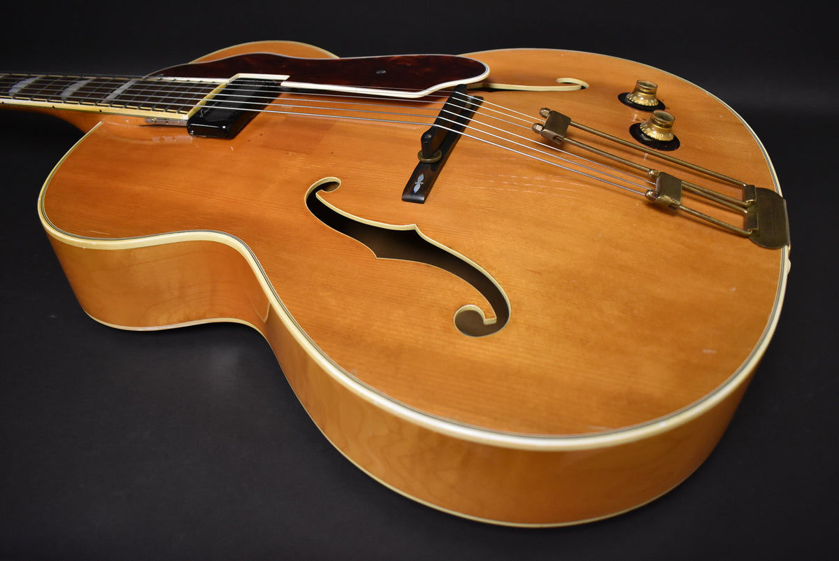 The natural finish archtop guitar, viewed from the upper bout, shows a dark pickguard, f-holes, and two gold control knobs.