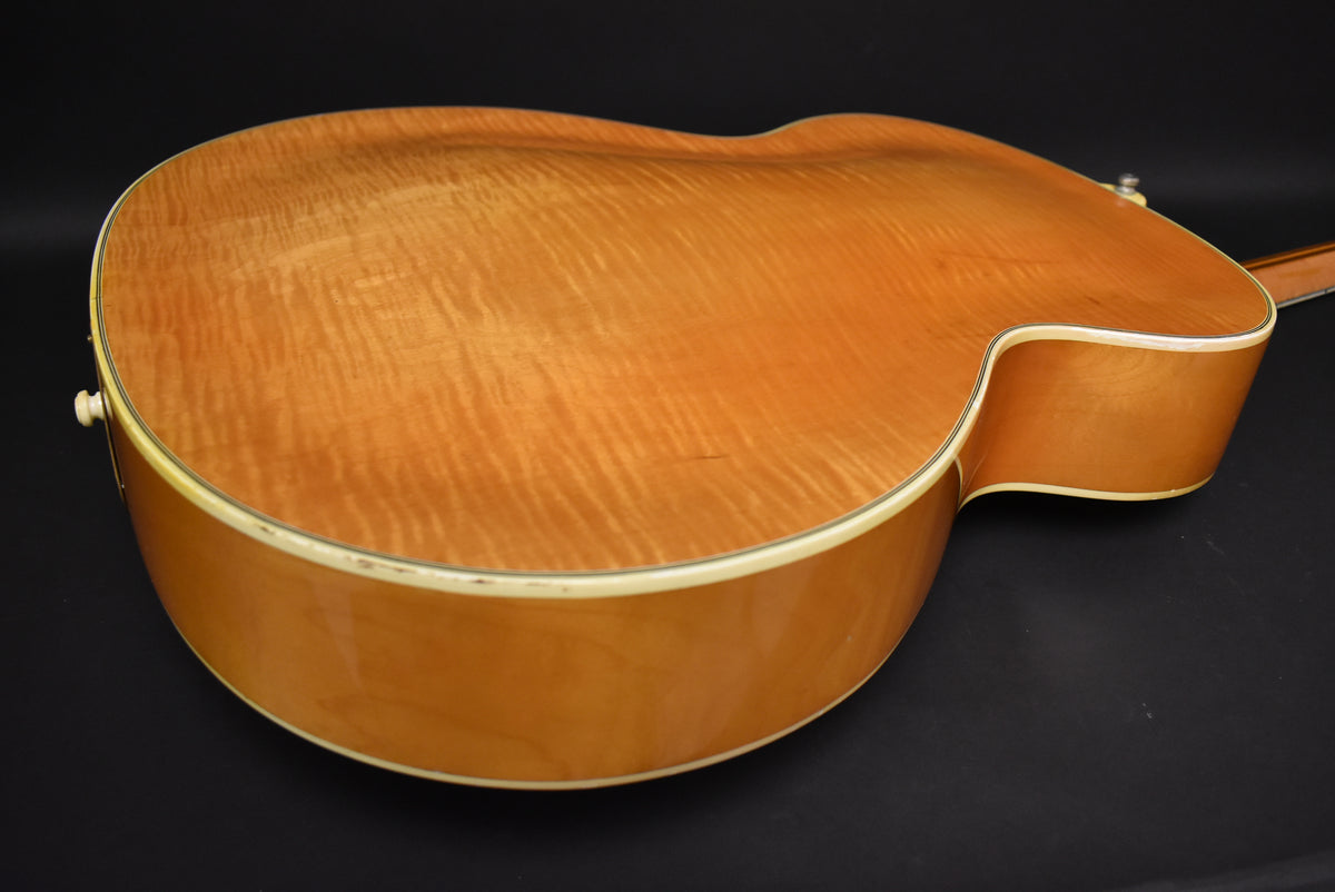 The archtop guitar's figured maple back and sides are visible from the lower bout, with binding wear and a strap button on the upper bout.