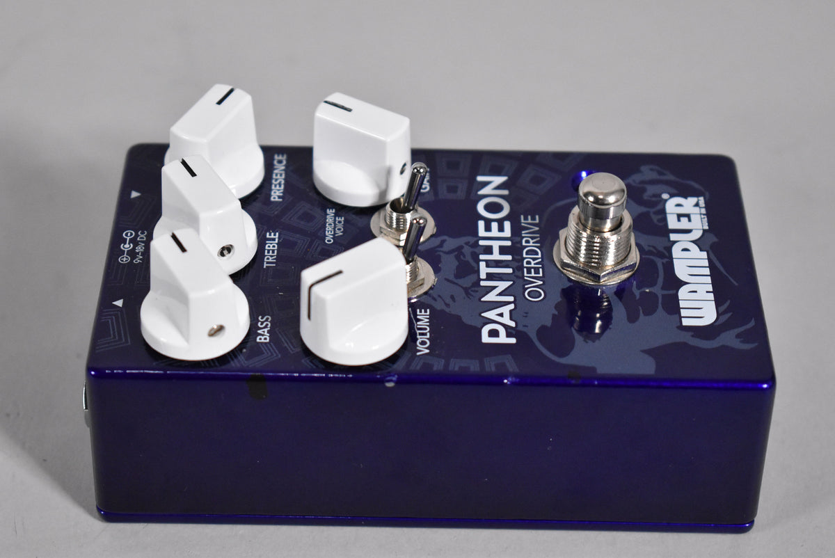 A dark purple pedal with white knobs and two small toggle switches, viewed from the top right, showing a silver footswitch.