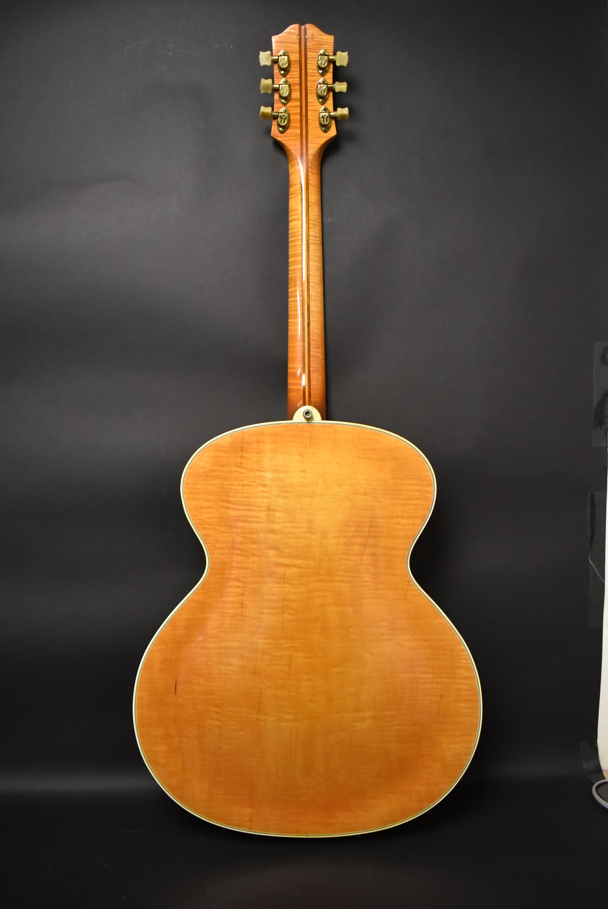 The guitar's back, neck, and headstock are visible, showing a natural finish with flame maple figuring and six gold tuners.