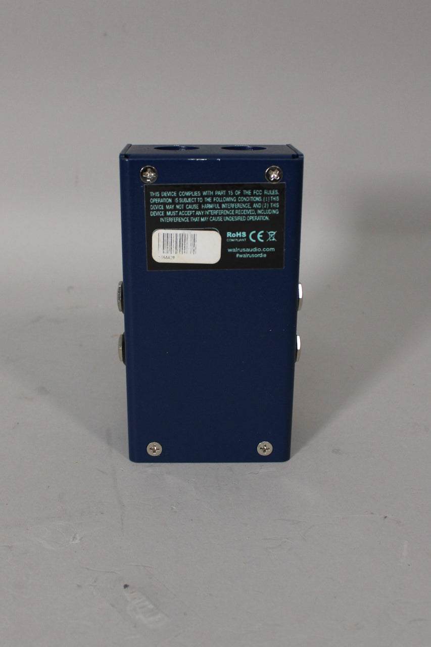 The dark blue pedal is viewed from the back, showing a black label with white text and a barcode, and four silver screws.
