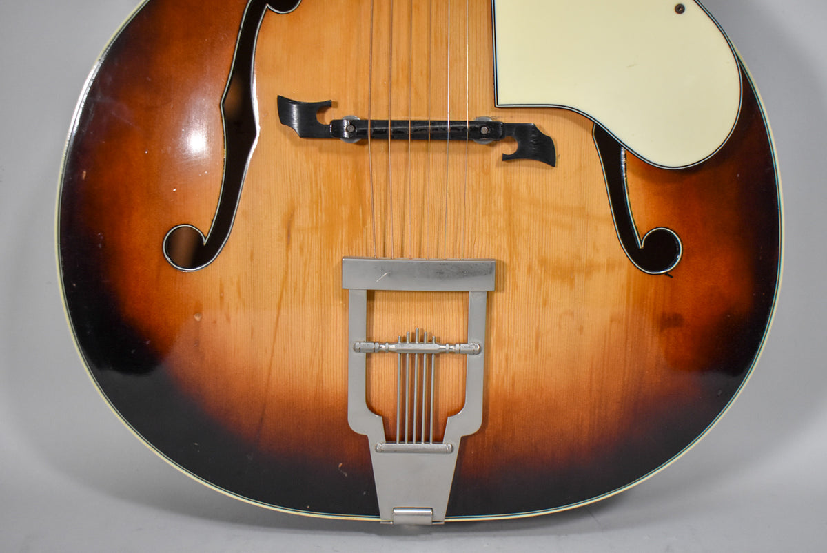Close-up of the sunburst archtop's lower body, showing the trapeze tailpiece, floating bridge, f-holes, and cream pickguard.